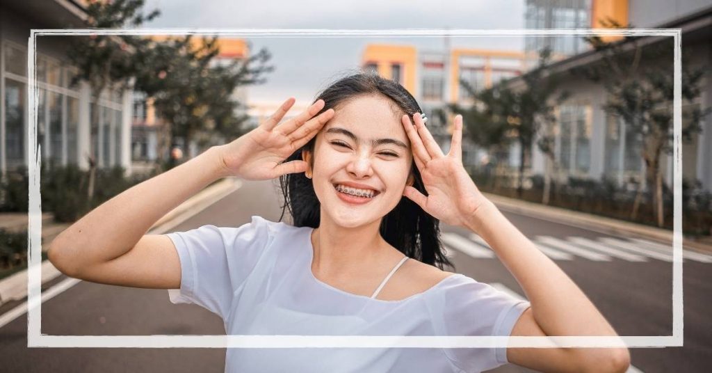 a smiling person with clear orthodontic braces posing hands near temples outdoors.