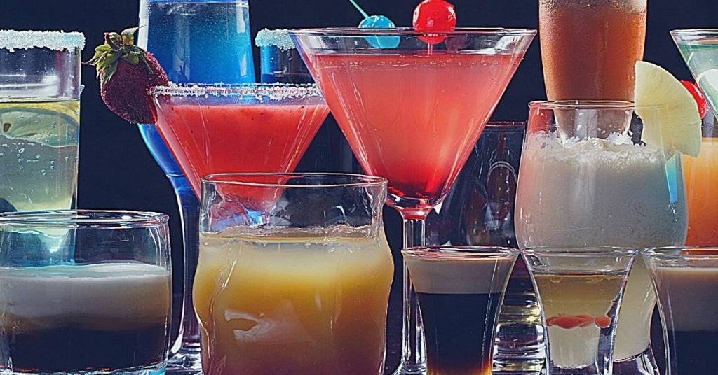 a colorful assortment of cocktails in martini and highball glasses, garnished with fruit and rimmed with salted sugar