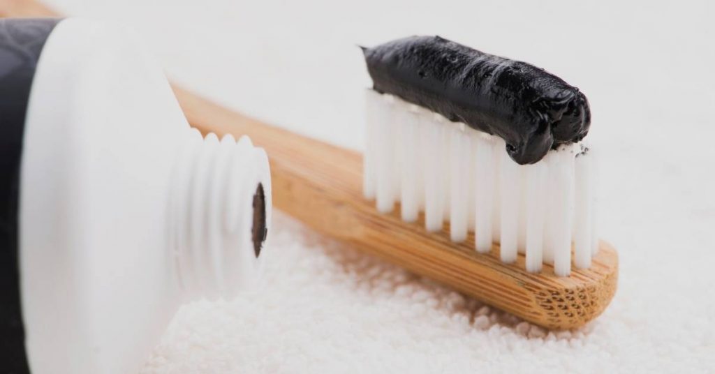 a wooden toothbrush holder with a bamboo toothbrush covered in black charcoal toothpaste residue, placed on a fluffy white surface.
