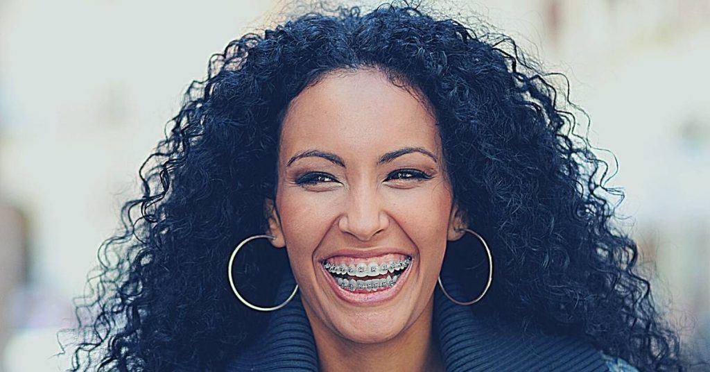 a smiling woman with curly hair wearing gold hoop earrings and clear dental braces