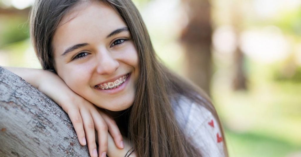 a smiling young person with braces leaning on a tree outdoors, showing confidence in orthodontic treatment