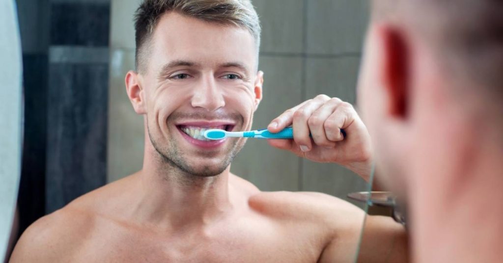 a smiling man brushing his teeth thoroughly with an electric toothbrush in a bathroom mirror.
