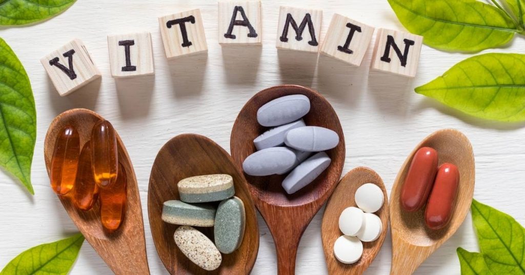 wooden spoons holding various vitamin and supplement capsules, with wooden blocks spelling "VITAMIN" and green leaves.