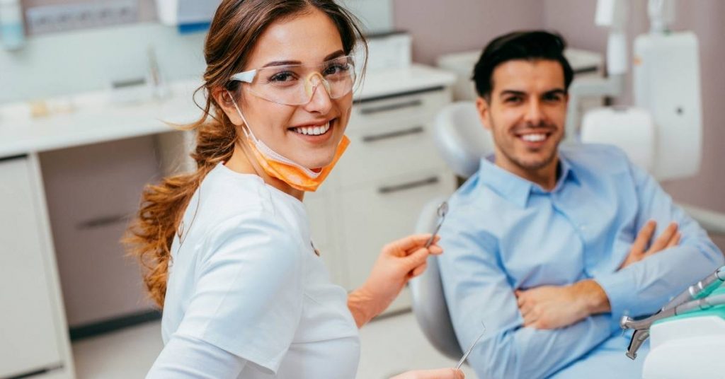 a dental professional in protective gear smiling while holding dental tools, demonstrating gum treatment techniques in a clinic setting