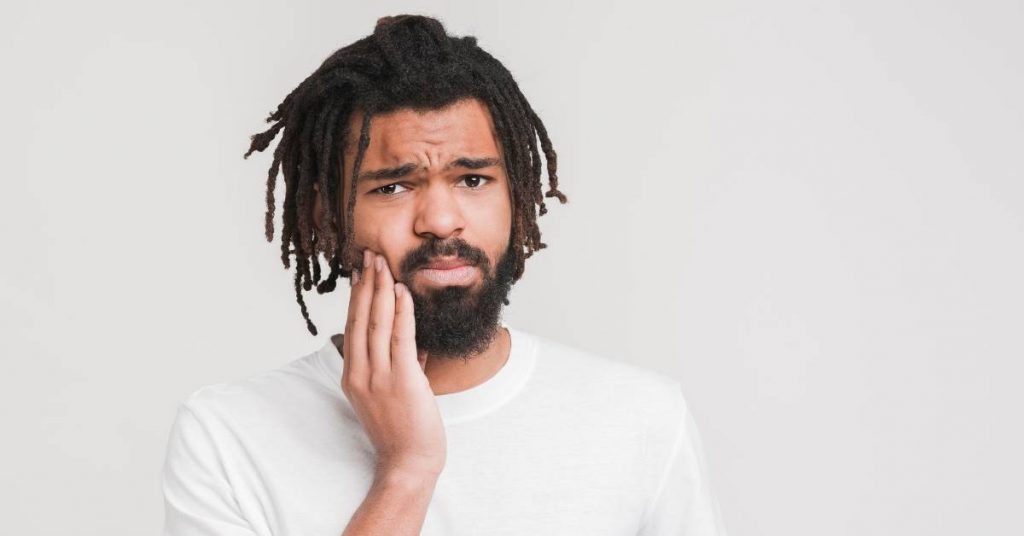 a person with dreadlocks holding their cheek in apparent distress from tooth pain