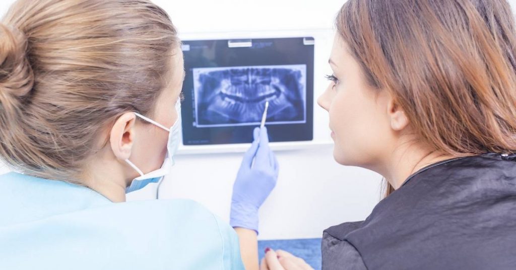 a dentist in protective gear pointing at a panoramic dental X-ray while discussing teeth and surrounding bone structure with a patient.