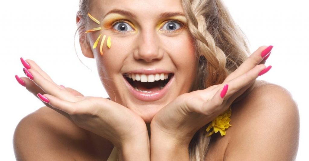a smiling person with bright pink nail polish holding hands to cheeks, showcasing wide-open mouth with gleaming teeth and yellow floral accents near eyes