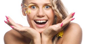 a smiling person with bright pink nail polish holding hands to cheeks, showcasing wide-open mouth with gleaming teeth and yellow floral accents near eyes