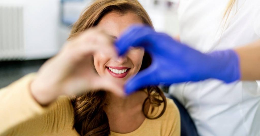 a smiling person making a heart shape with their hands, wearing a yellow shirt, post-recovery from oral surgery.