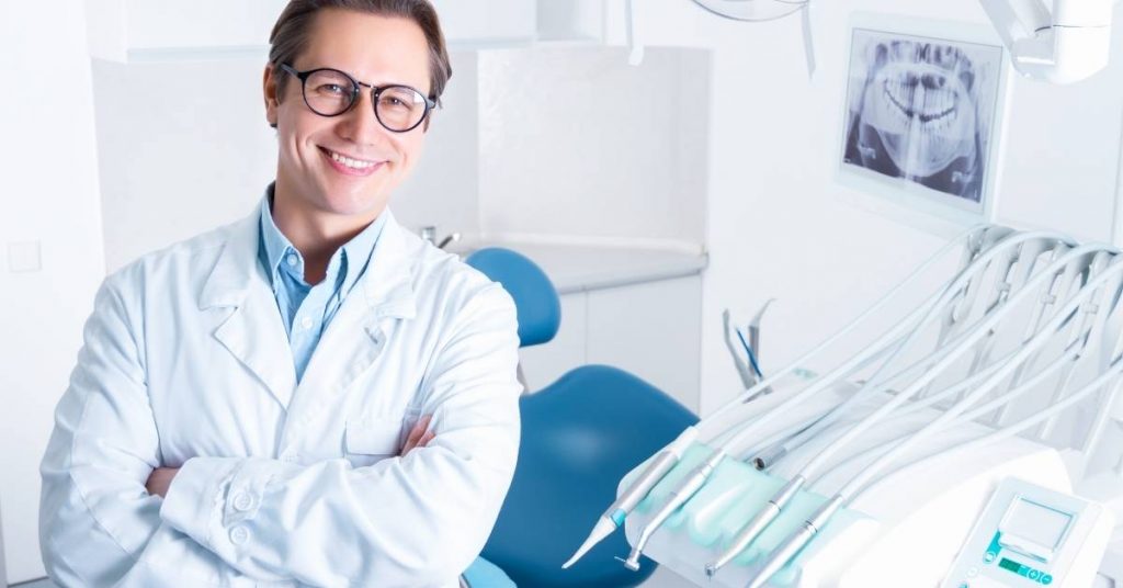 a smiling dentist in a clinic wearing a white lab coat, standing confidently with dental tools and an X-ray of teeth in the background