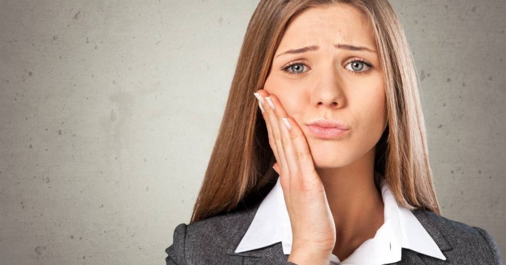a woman in professional attire holding her jaw in discomfort, suggesting potential oral pain or dental issue