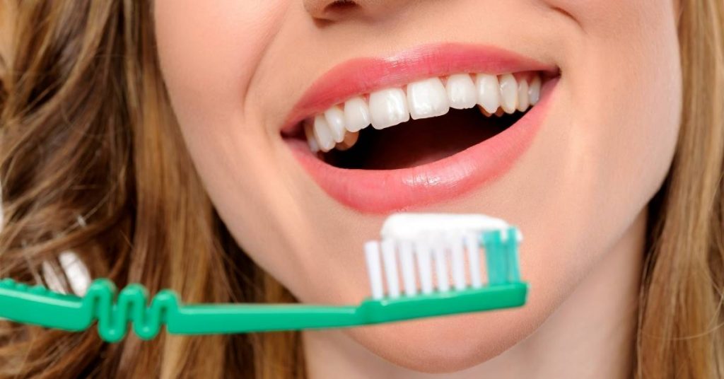 a person smiling widely showing healthy teeth while holding a green toothbrush, emphasizing proper oral hygiene for gum health.