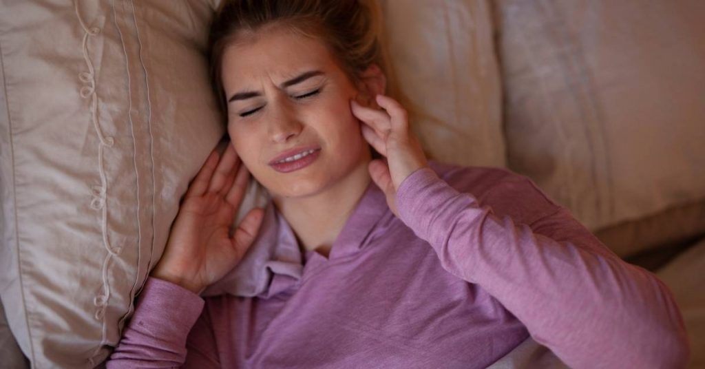 a person lying in bed holding their ear, showing signs of jaw muscle tension or facial pain.