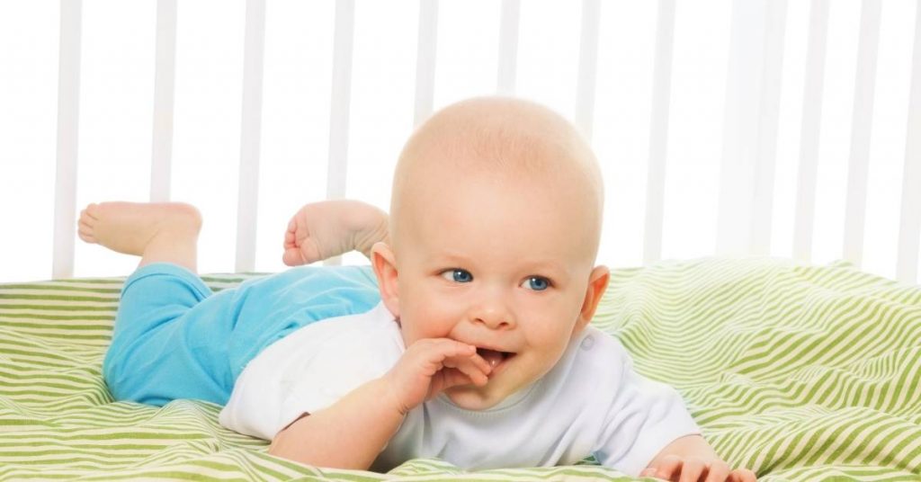 a baby lying on a textured blanket, touching their mouth with one hand while looking curious