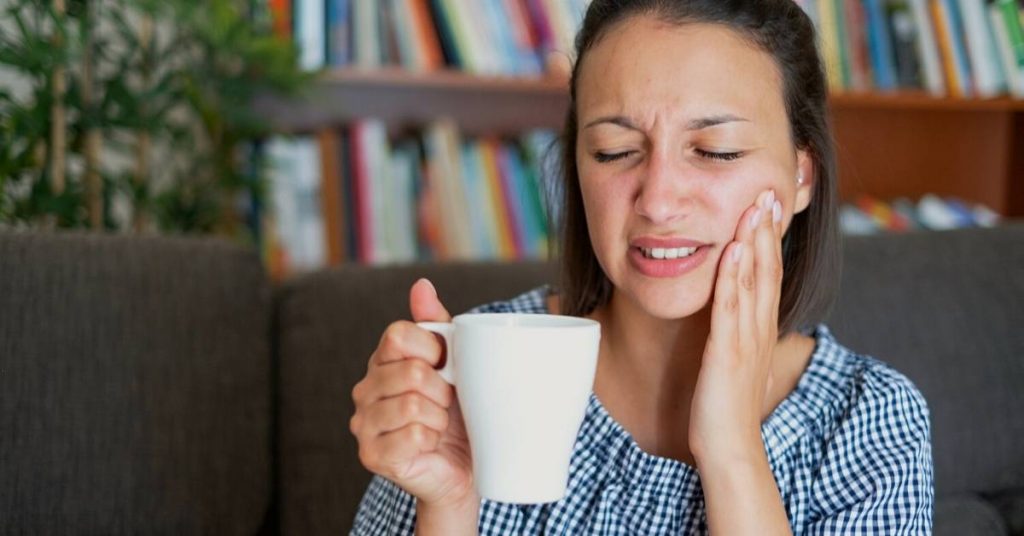 a person holding a cup while touching their cheek, indicating discomfort or sensitivity possibly related to teeth root exposure.