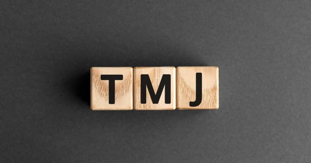 three wooden block letters spelling "TMJ" against a dark background