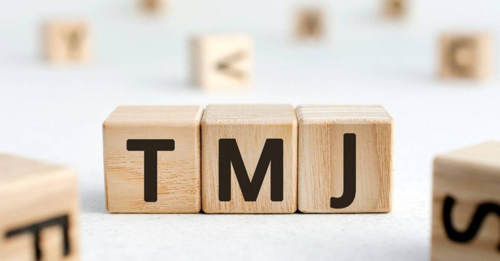 wooden block letters spelling "TMJ" surrounded by scattered alphabet and number blocks on a light background