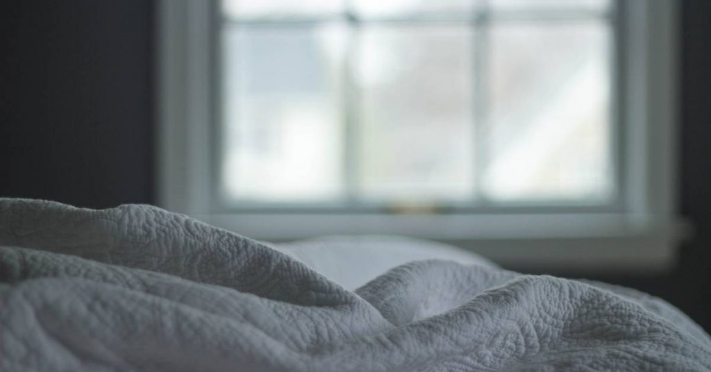 a neatly made bed with soft, rumpled gray bedding viewed from the foot, looking toward a large window with natural light.