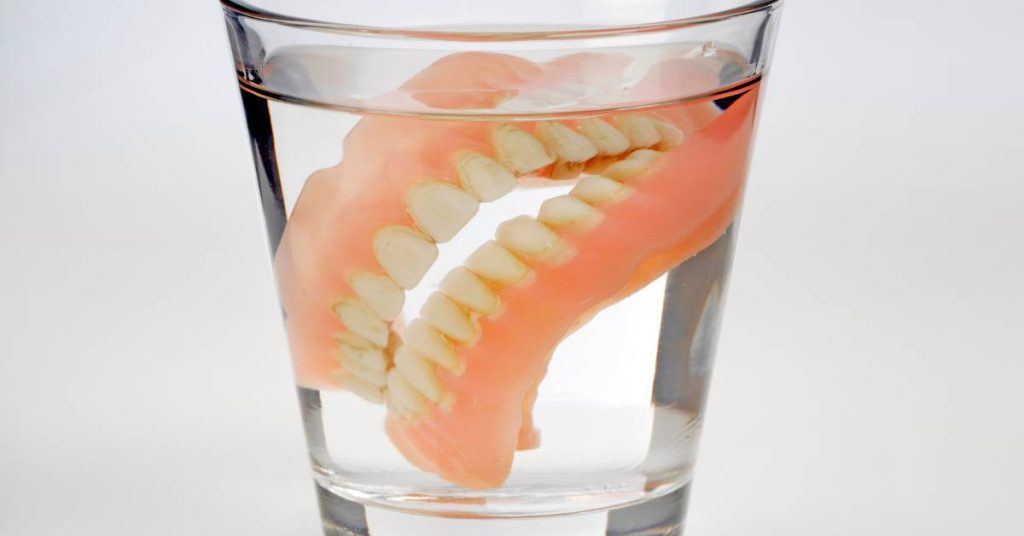 a clear glass filled with water holding a pink plastic model of lower dentures for visualizing replacing dentures