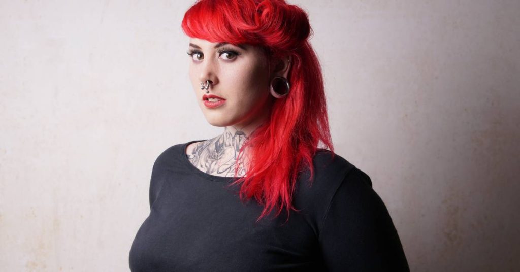 a person with vibrant red hair styled in a high ponytail, featuring a nose piercing and ear piercings, showcasing visible facial tattoos and piercings.