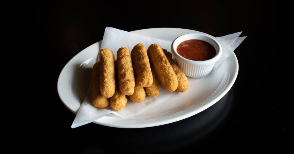 a white plate with six golden-fried mozzarella sticks on parchment, paired with a small cup of marinara sauce