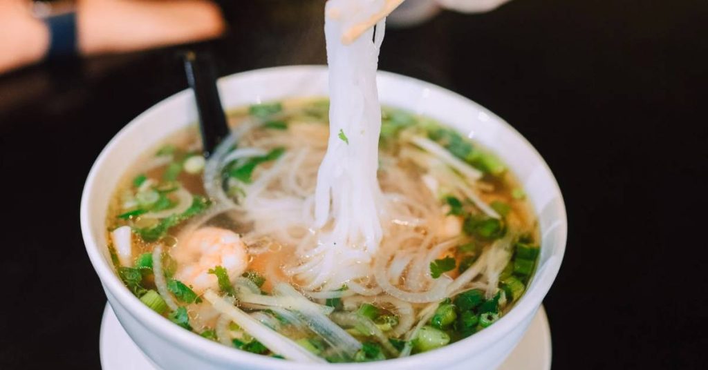 a bowl of Vietnamese pho with soft rice noodles, shrimp, and green onions being lifted with chopsticks