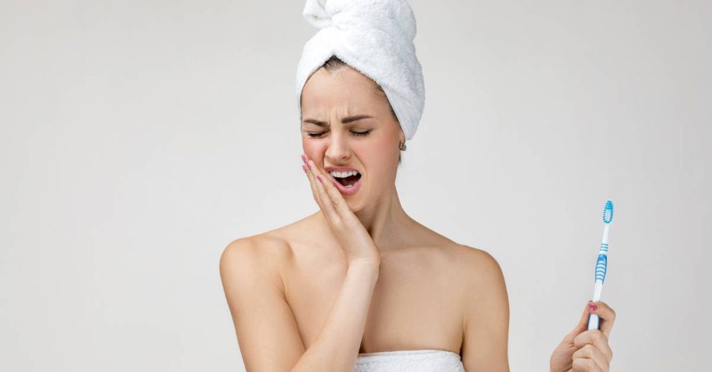 a person in a towel turban holding a toothbrush while grimacing in pain near their extracted wisdom teeth area