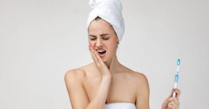 a person in a towel turban holding a toothbrush while grimacing in pain near their extracted wisdom teeth area