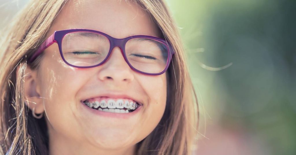 a smiling young person with braces on their teeth and purple-rimmed glasses outdoors