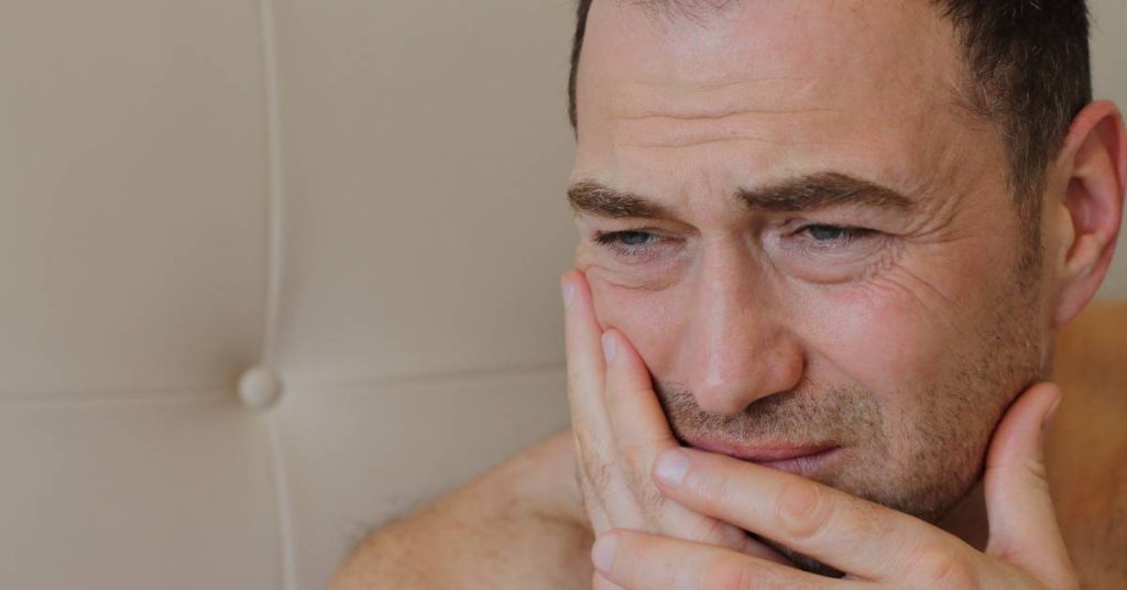 a man holding his hand to his chin, appearing to experience tooth pain or sensitivity while indoors.