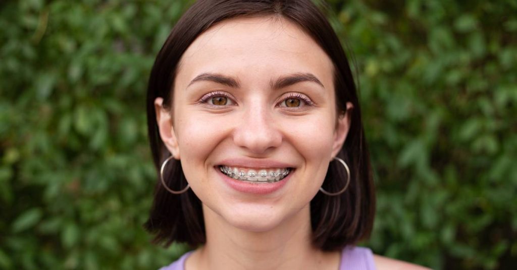 a smiling person wearing clear braces and hoop earrings against a green foliage background