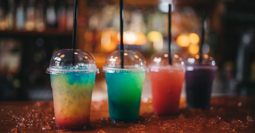four colorful slushie drinks in transparent cups with straws, placed on crushed ice